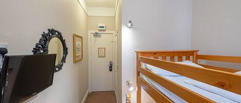 Une chambre avec un lit superposé, un téléviseur à écran plat fixé au mur, une table en bois et une porte avec un interrupteur et une petite pancarte au-dessus.