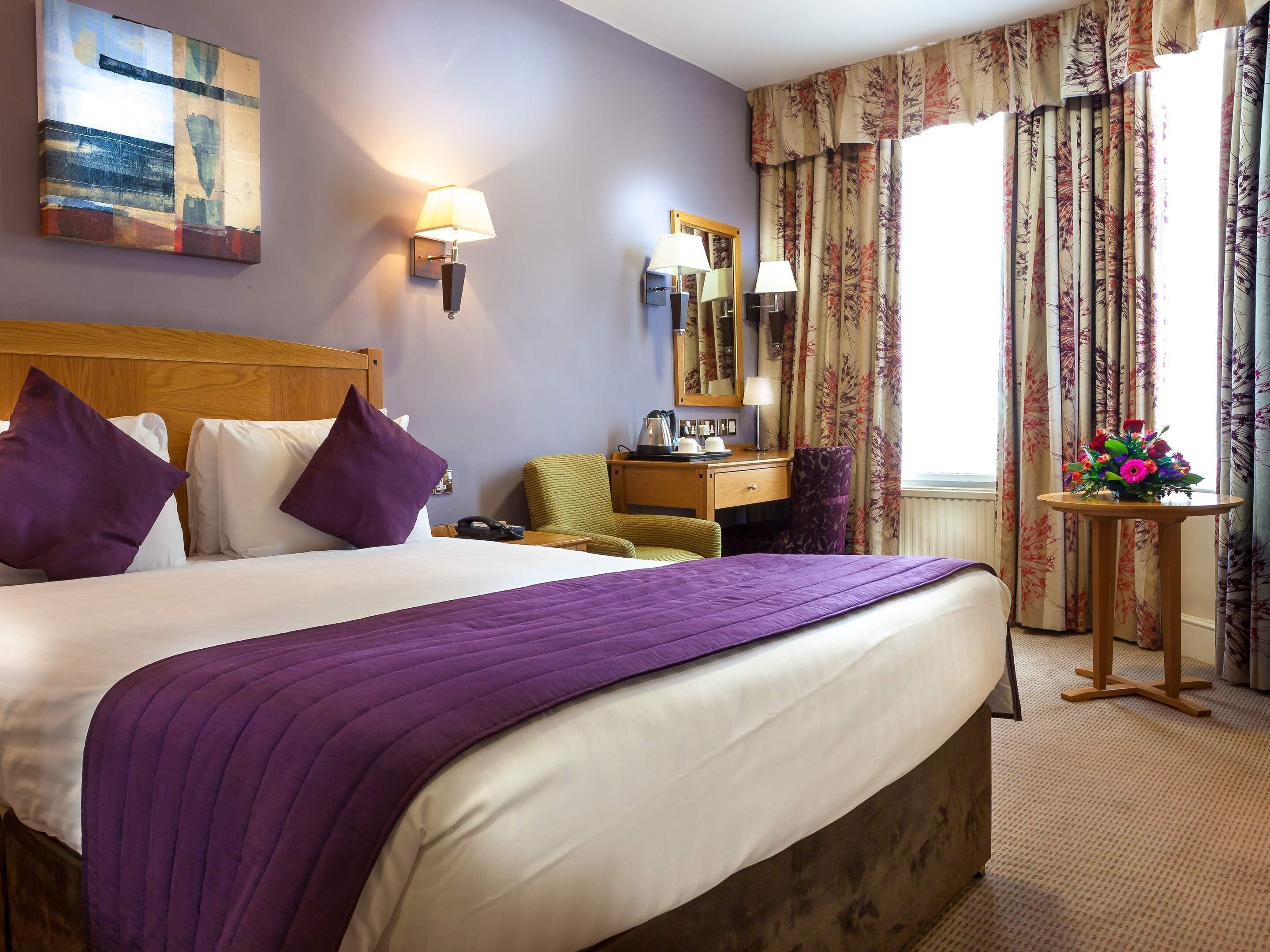 A hotel room with a large bed, a wooden headboard, a desk with a chair, a small table with a vase of flowers, and a window with curtains.