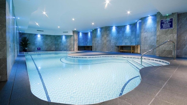 A modern indoor swimming pool with a curved design, surrounded by stone walls and blue lighting. - The Durley Dean Hotel (Bournemouth)