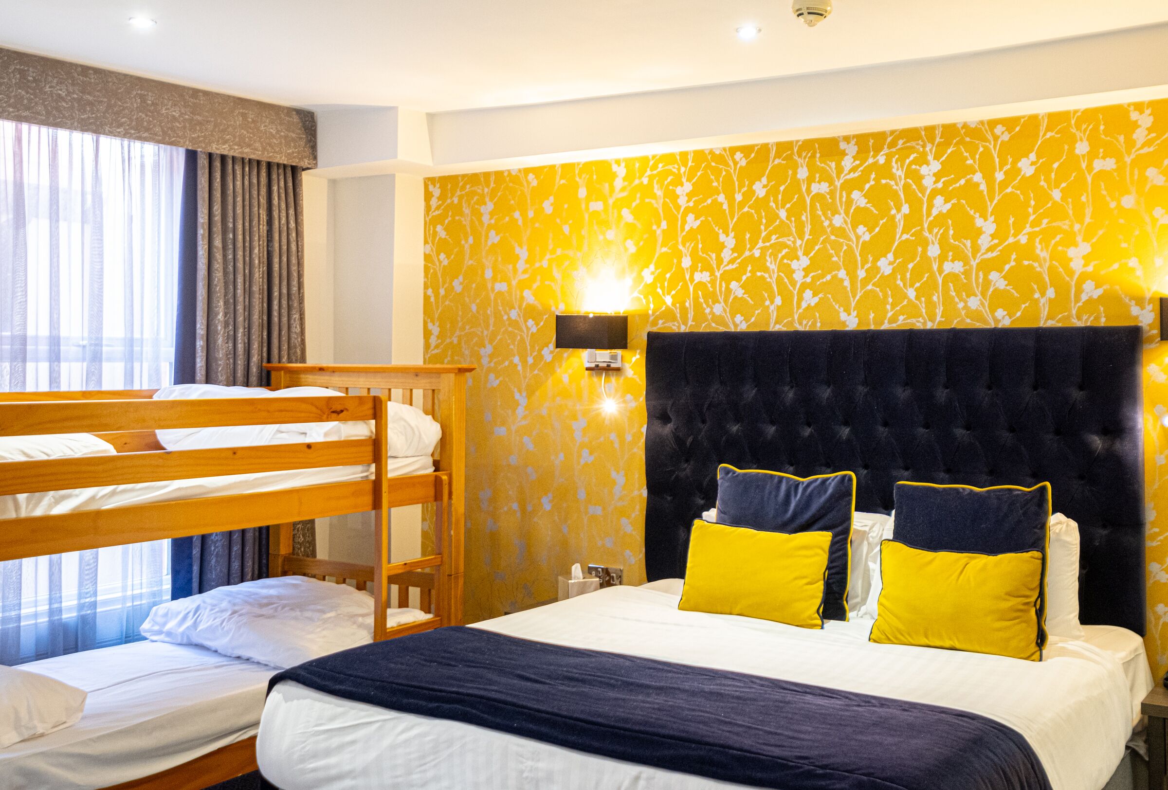 A hotel room with a bunk bed, a large bed with yellow and blue pillows, a wall-mounted lamp, and a window with curtains.
