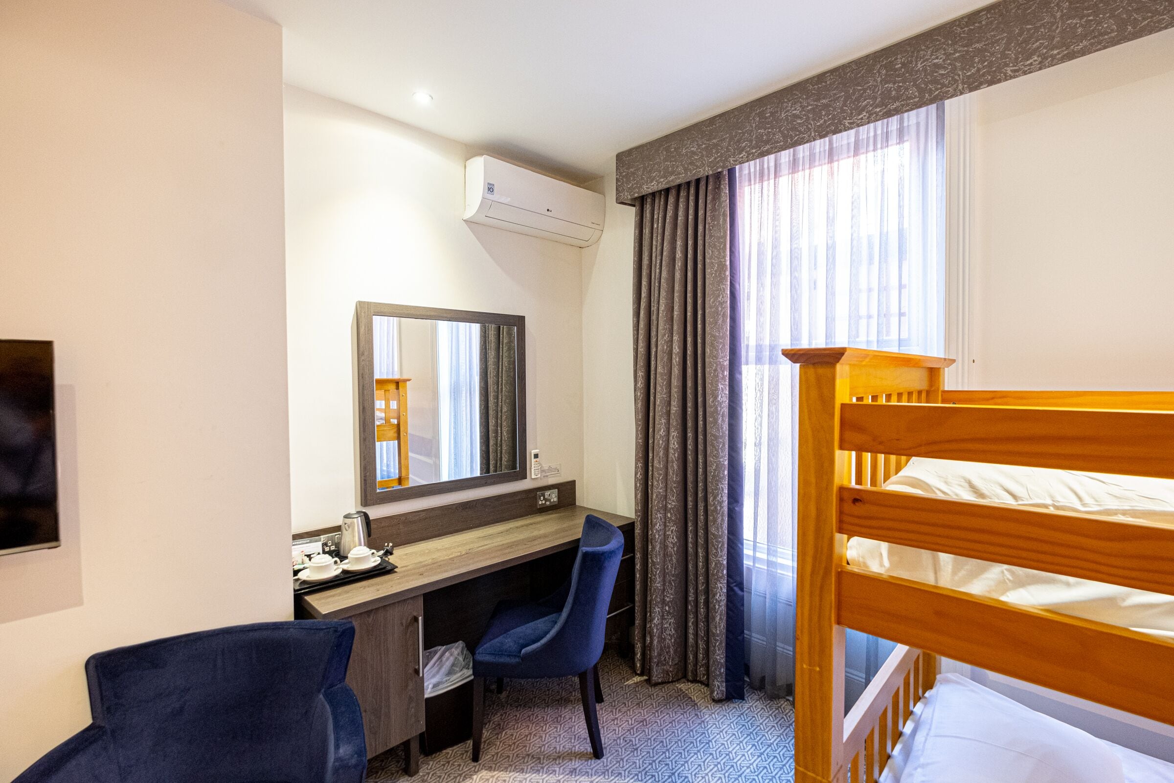 A hotel room with a bunk bed, a desk with a chair, a large mirror, and a window with curtains.