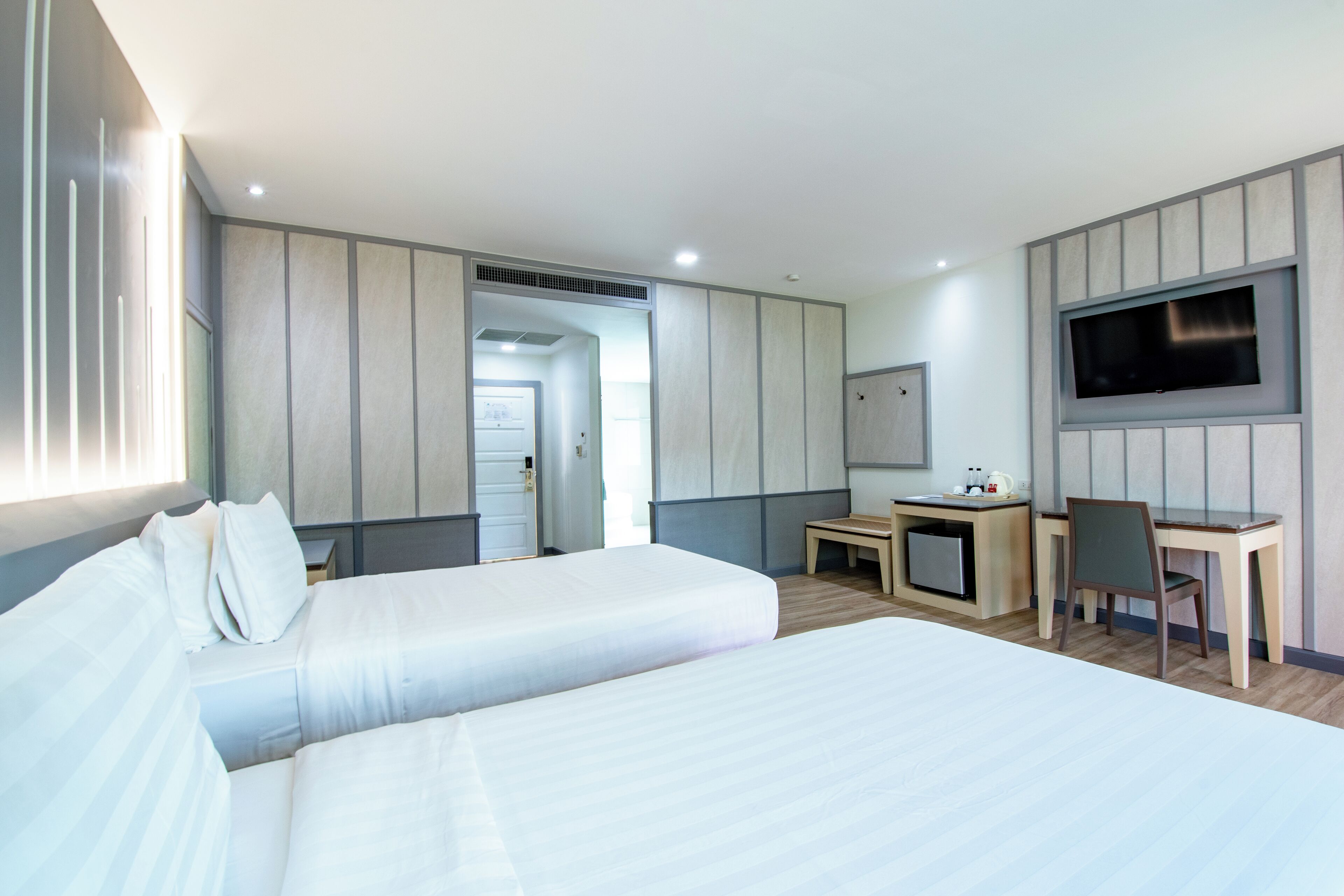 A hotel room with two beds, a desk, a chair, a TV mounted on the wall, and a small kitchenette.