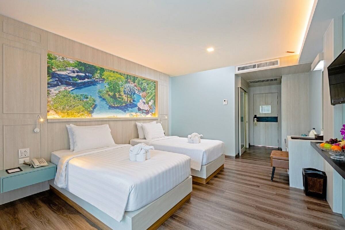 A hotel room with two beds, a painting of a landscape on the wall, a desk with a computer, and a small kitchenette.