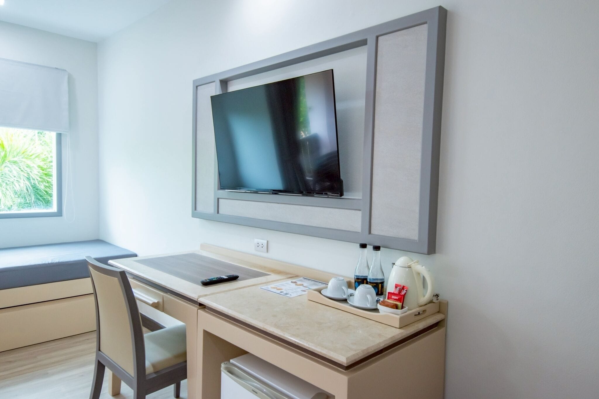 A hotel room with a bed, a desk with a chair, a television mounted on the wall, and a window with a view of greenery.
