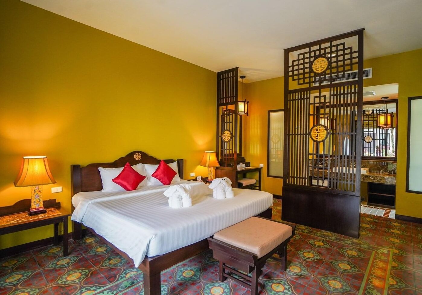 A hotel room with a bed, two bedside tables, a bench, a decorative screen, and a tiled floor.