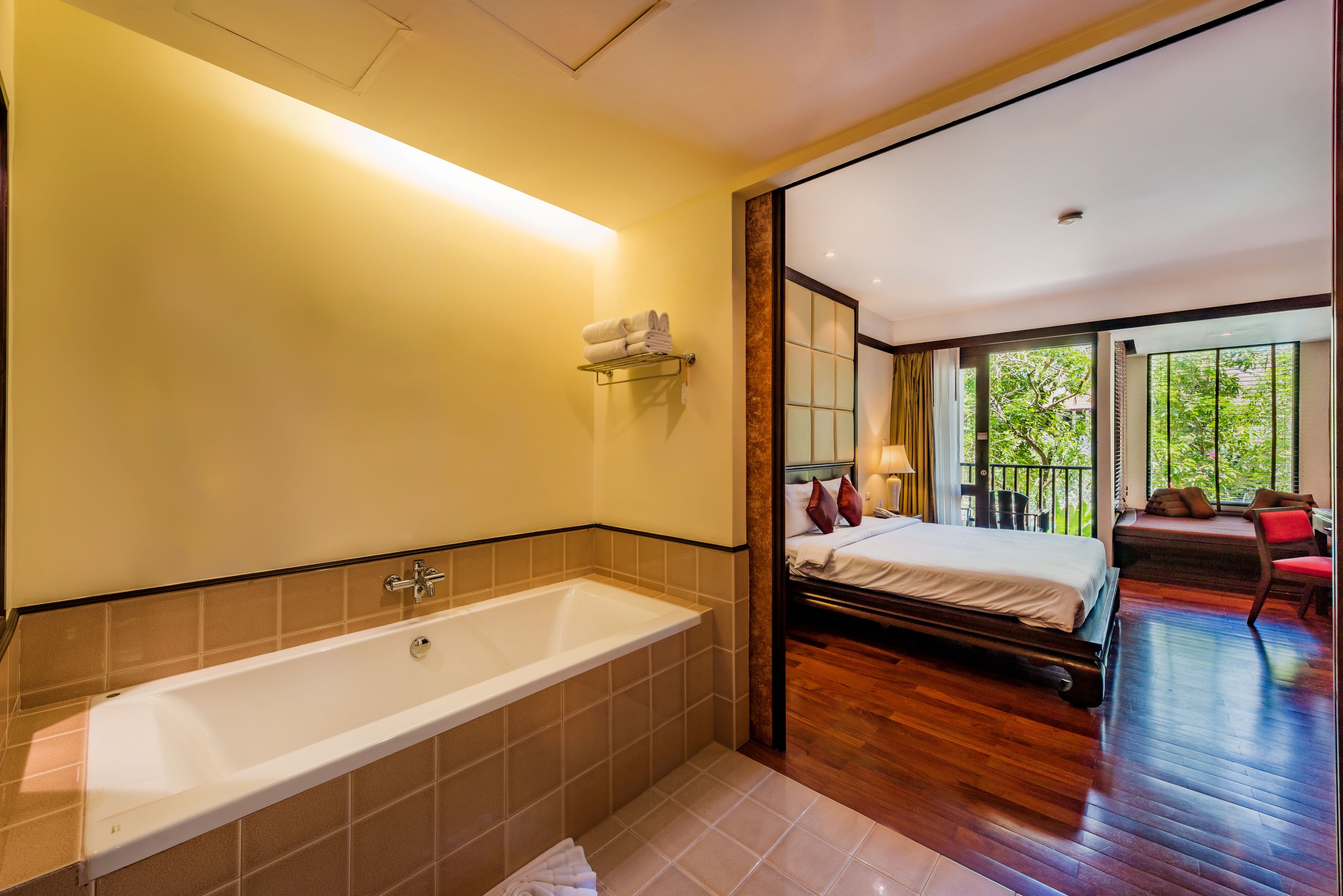 A modern bathroom with a bathtub, a shower, and a towel rack. The room has a wooden floor and a large window offering a view of greenery outside.