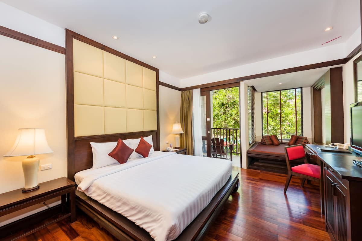 A hotel room with a large bed, a desk with a chair, and a balcony with a view of greenery.