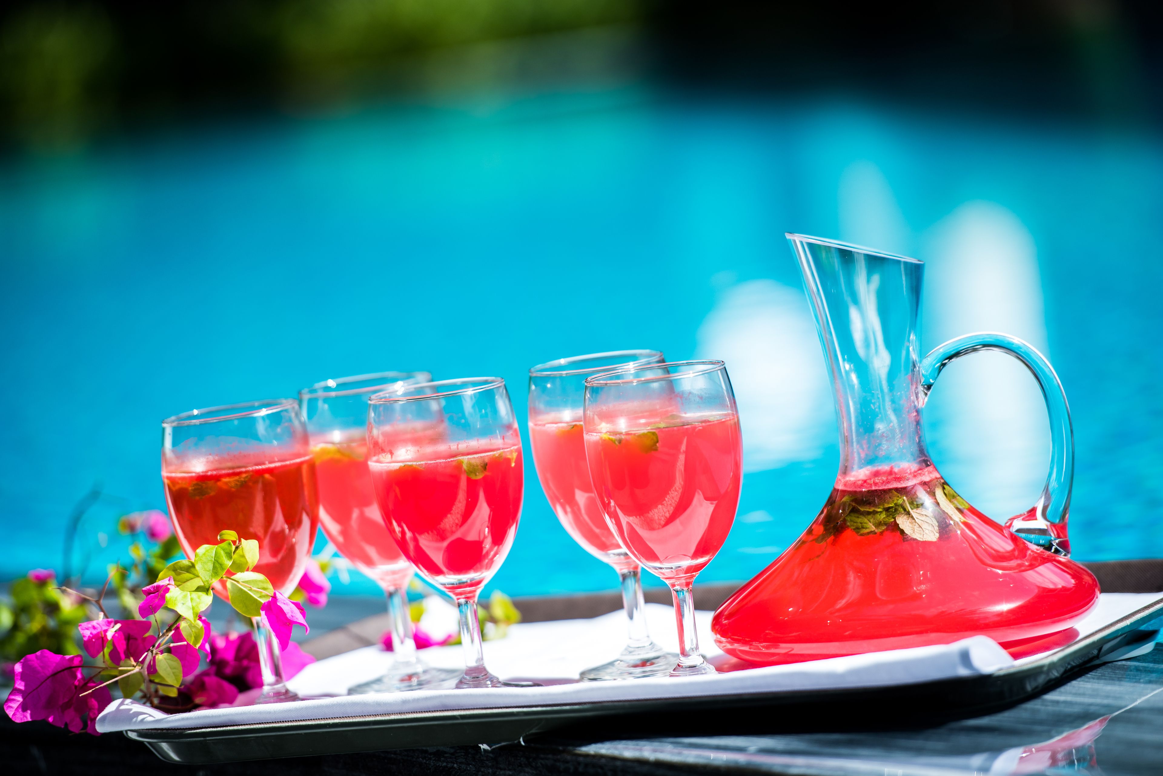 A tray with four glasses and a pitcher of red drink, placed on a table with a pool in the background.