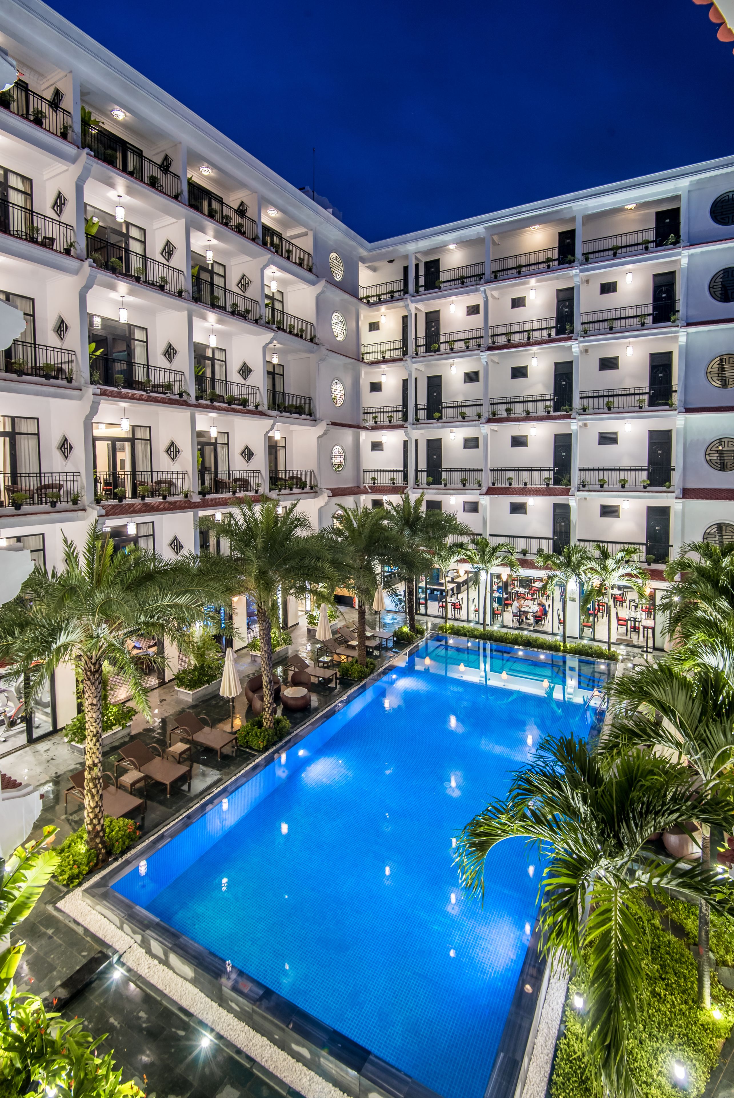A multi-story hotel with a central swimming pool surrounded by palm trees and outdoor seating areas.