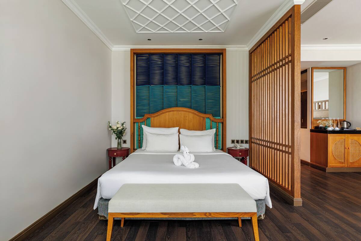 A neatly made bed with a wooden headboard, a bedside table with a vase of flowers, and a wooden partition with a woven design.