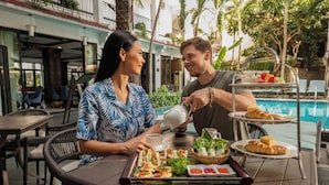 A dining table with a tray of food and drinks, a pool in the background, and a building with balconies. - Hadana Boutique Resort Hoi An (Da Nang)