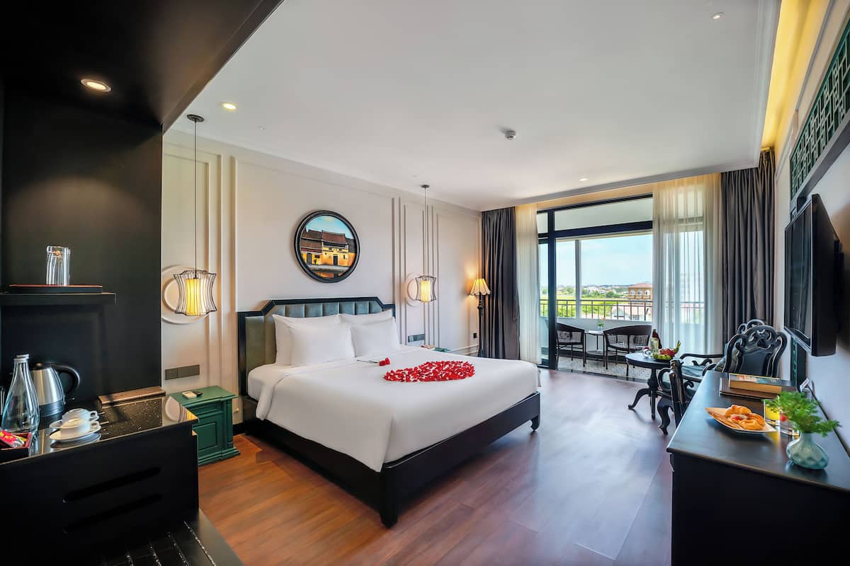 A hotel room with a large bed, a desk with a chair, a dining table, and a view of the city through large windows.