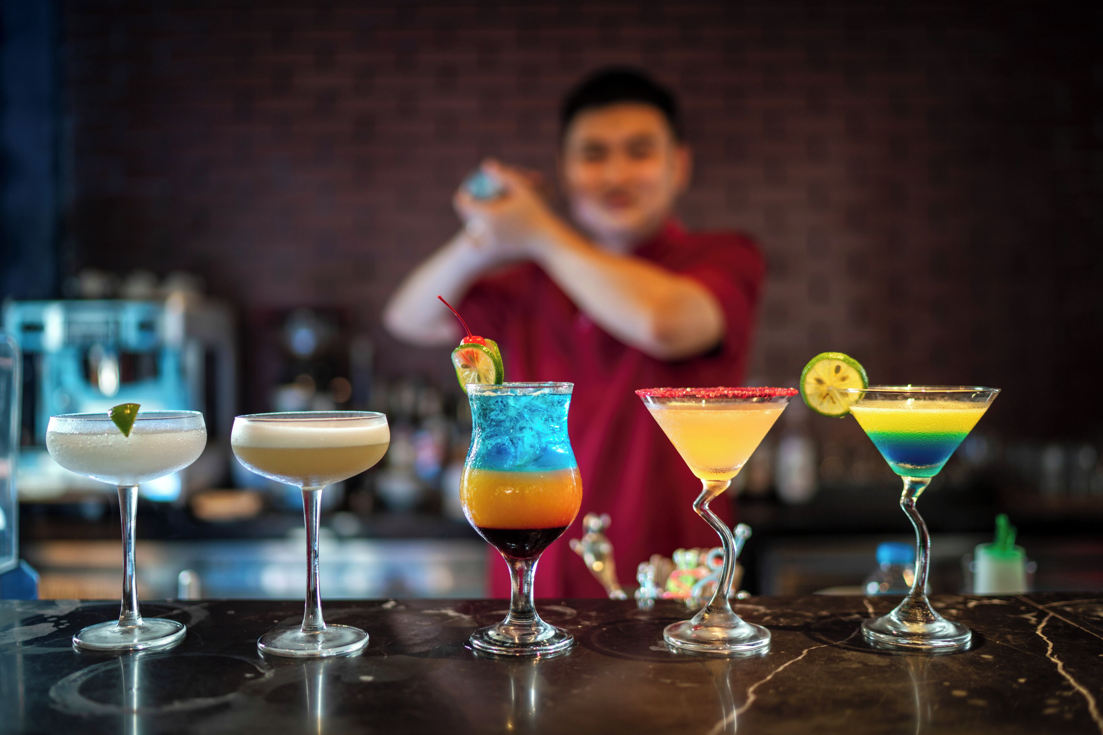 A bar with four colorful cocktails and a bartender preparing drinks.