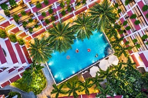 An aerial view of a hotel with a central swimming pool surrounded by palm trees and a red-tiled walkway.