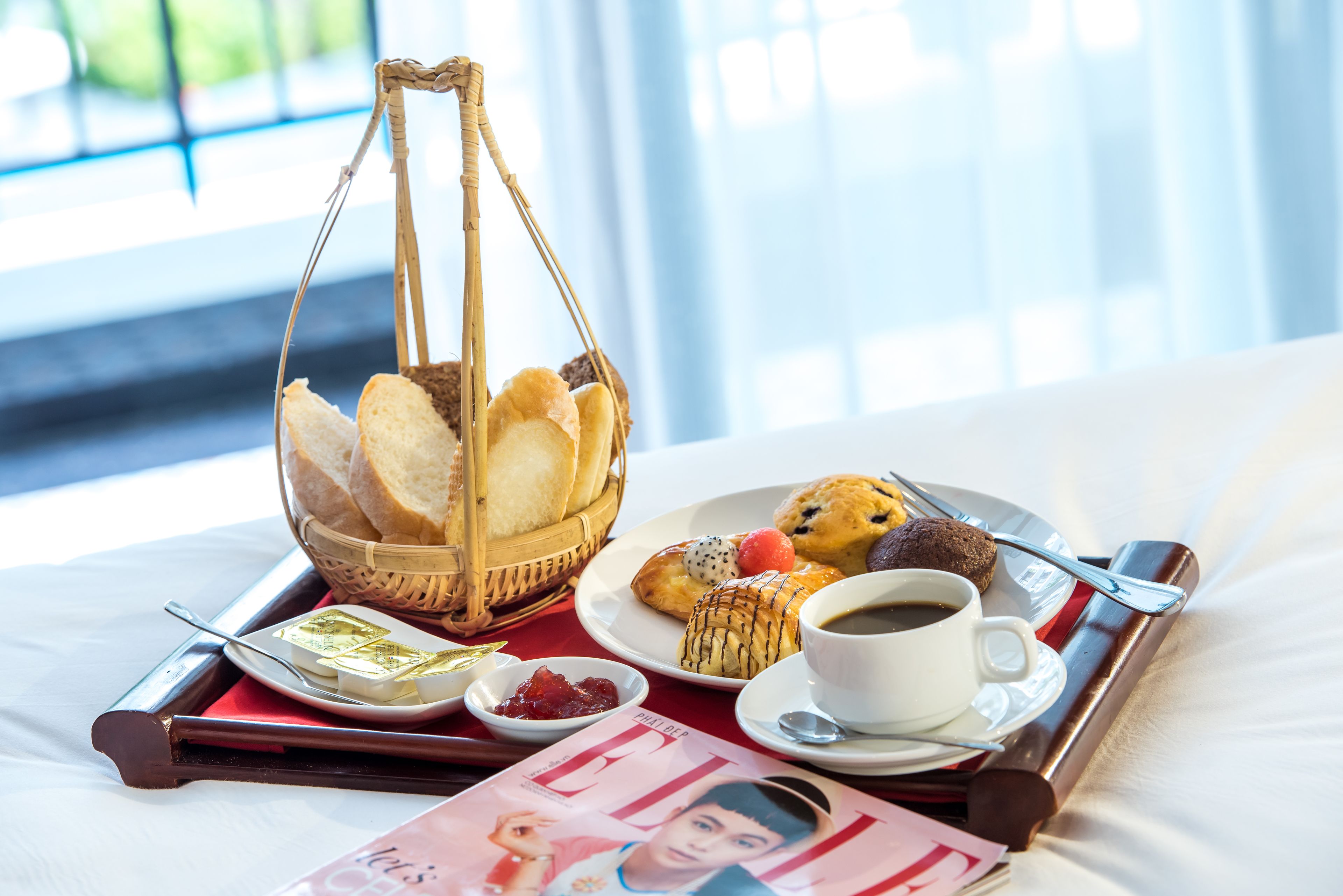 A breakfast tray with bread, pastries, coffee, and condiments on a bed with a magazine.