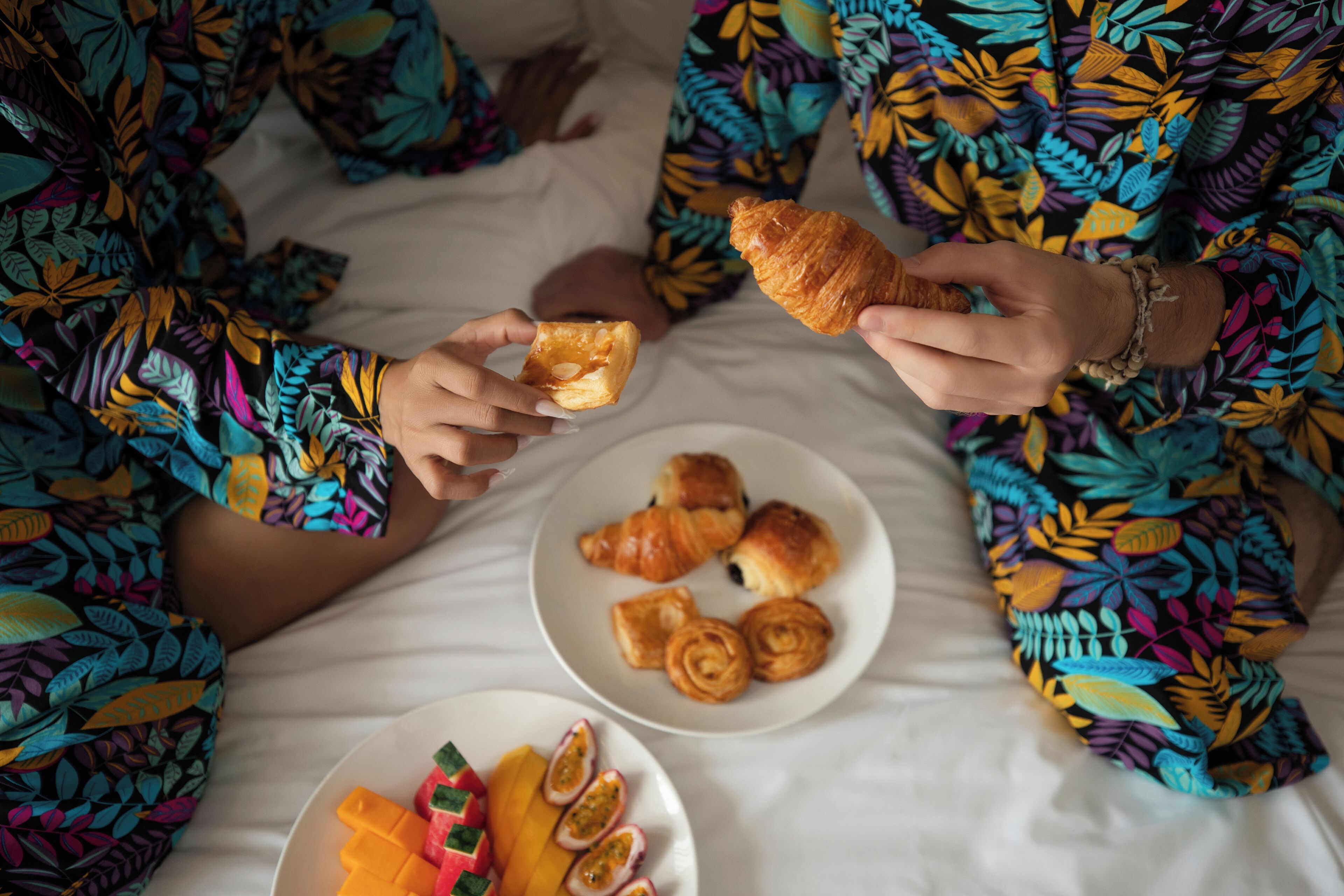 Deux personnes, vêtues de robes colorées à motifs, sont assises sur un lit devant des assiettes remplies de pâtisseries et de fruits.