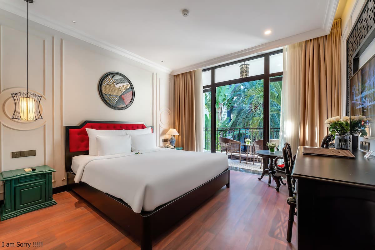 A hotel room with a large bed, a desk, a chair, a small table, a lamp, and a view of greenery outside.