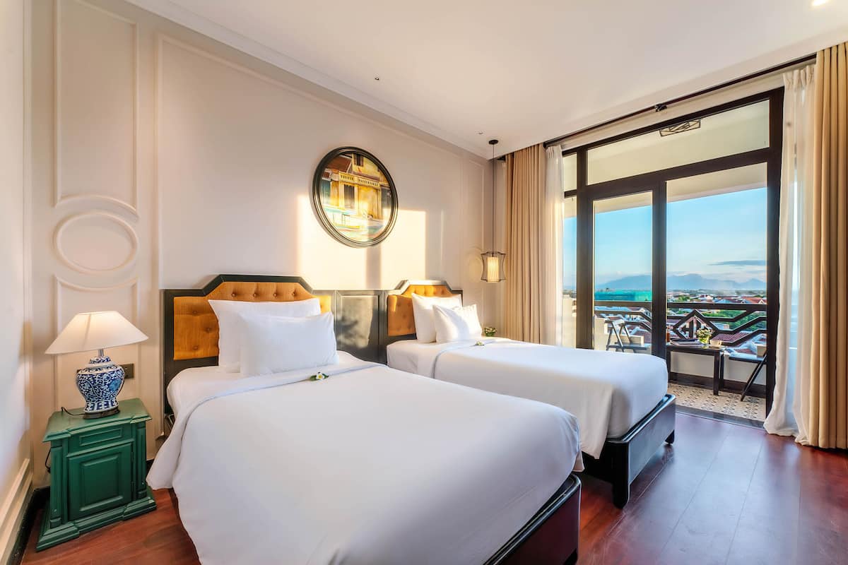 A hotel room with two beds, a large window with a view, a round mirror on the wall, and a green nightstand with a lamp.