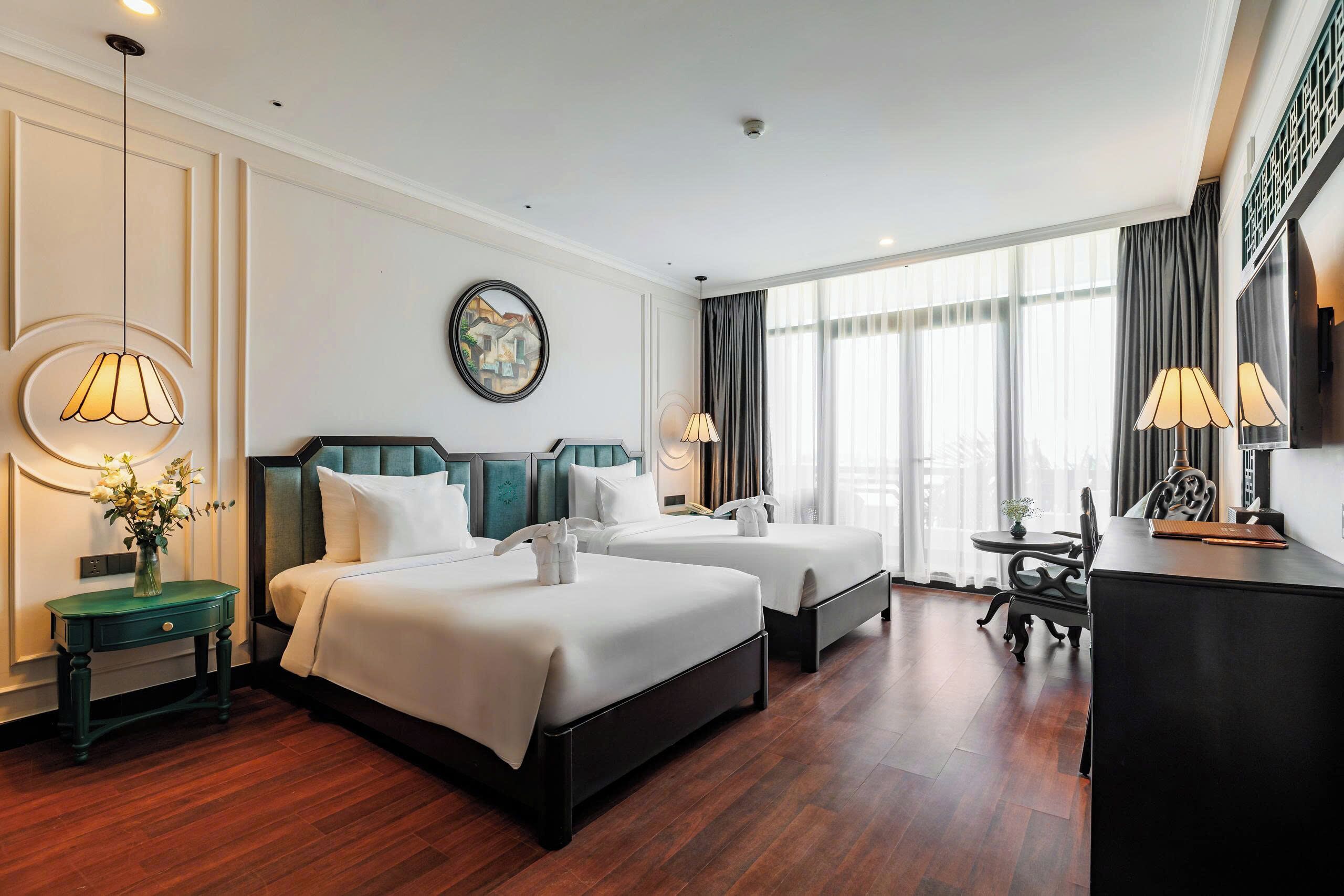 A hotel room with two beds, a desk, a chair, a vase with flowers, and a large window with curtains.