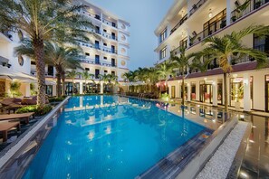 A large indoor swimming pool surrounded by palm trees and lounge areas, with multiple floors of hotel buildings in the background. - Hadana Boutique Resort Hoi An (Da Nang)