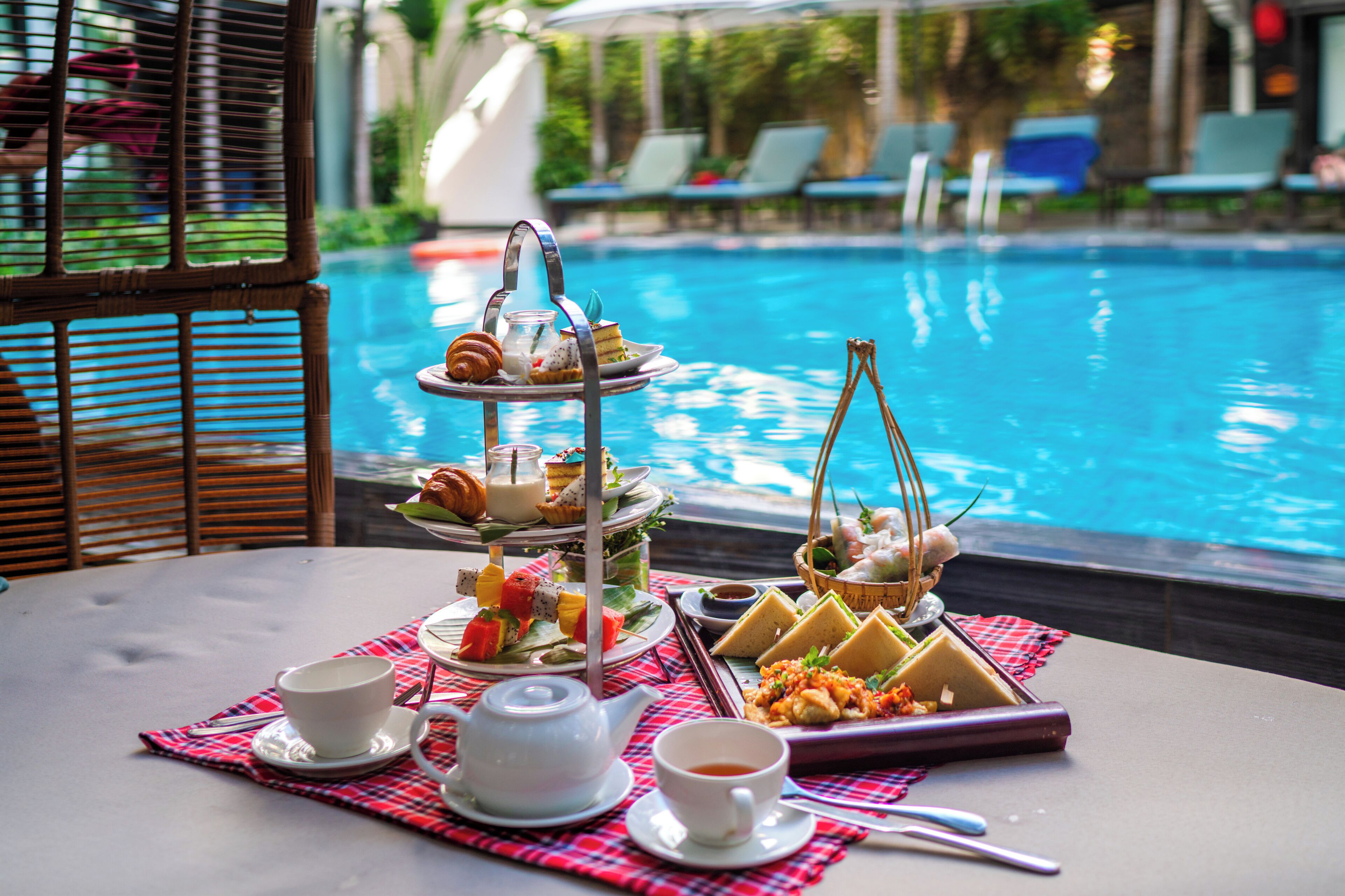 A table with a three-tiered dessert stand, a teapot, cups, and a basket of food by a poolside.