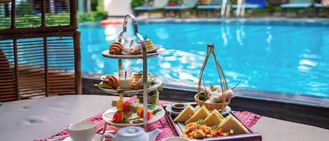 A table with a three-tiered dessert stand, a teapot, cups, and a basket of food by a poolside.
