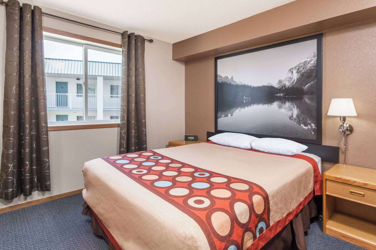 Ett hotellrum med en stor säng, ett nattduksbord, en lampa och ett fönster med gardiner.