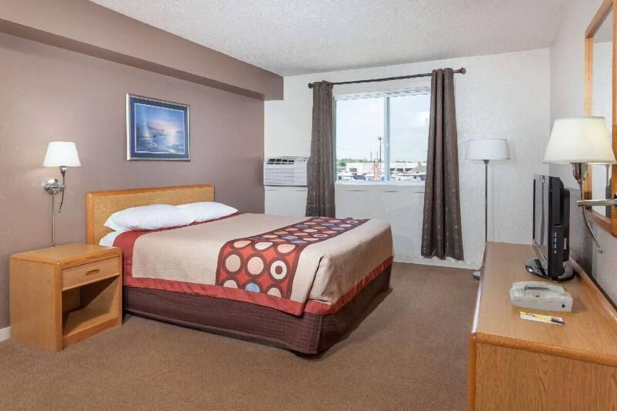 Ett hotellrum med en säng, ett skrivbord, en tv, en lampa, en tavla på väggen och ett fönster med gardiner.