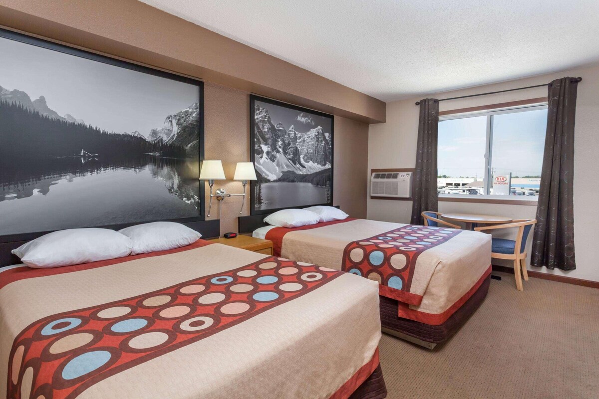 A hotel room with two beds, a desk, a chair, a window with curtains, and two wall-mounted paintings.