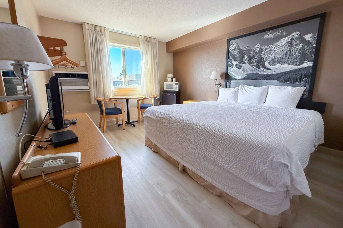 Ett hotellrum med en stor säng, ett skrivbord med en telefon, en stol, ett litet bord, en tv och ett fönster med gardiner.