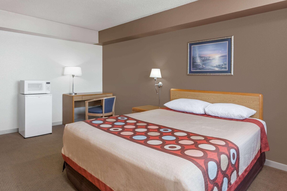 A hotel room with a bed, a desk with a chair, a microwave, two lamps, and a painting on the wall.