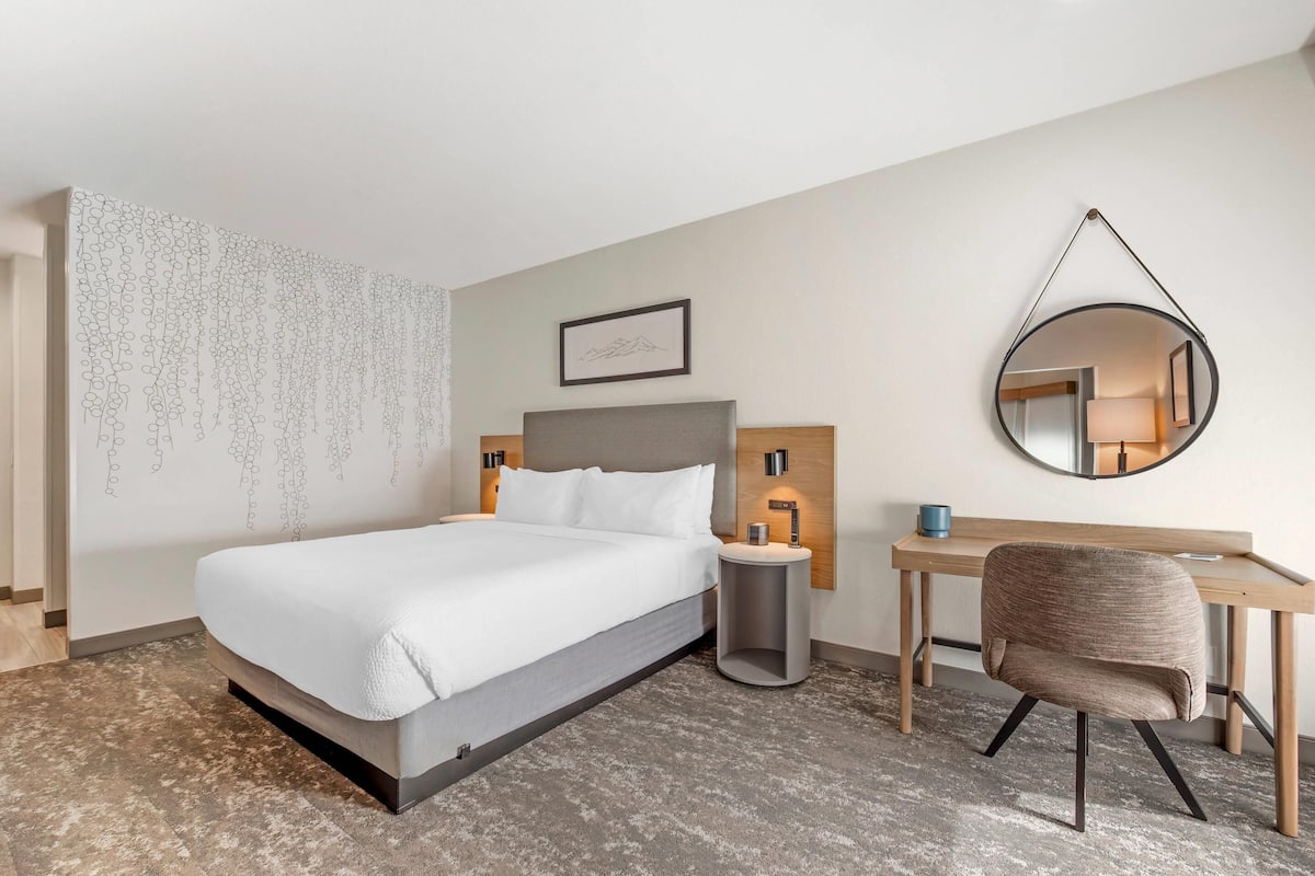 A modern hotel room with a bed, desk, chair, and a wall-mounted mirror.