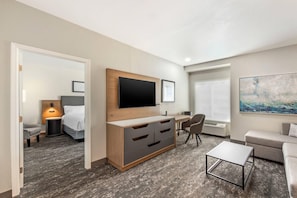 Una habitación de hotel moderna con una televisión de pantalla plana montada en un mueble de madera, un escritorio con una silla, un sofá y una pequeña mesa de centro.