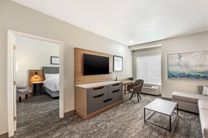 A modern hotel room with a flat-screen TV mounted on a wooden cabinet, a desk with a chair, a sofa, and a small coffee table.