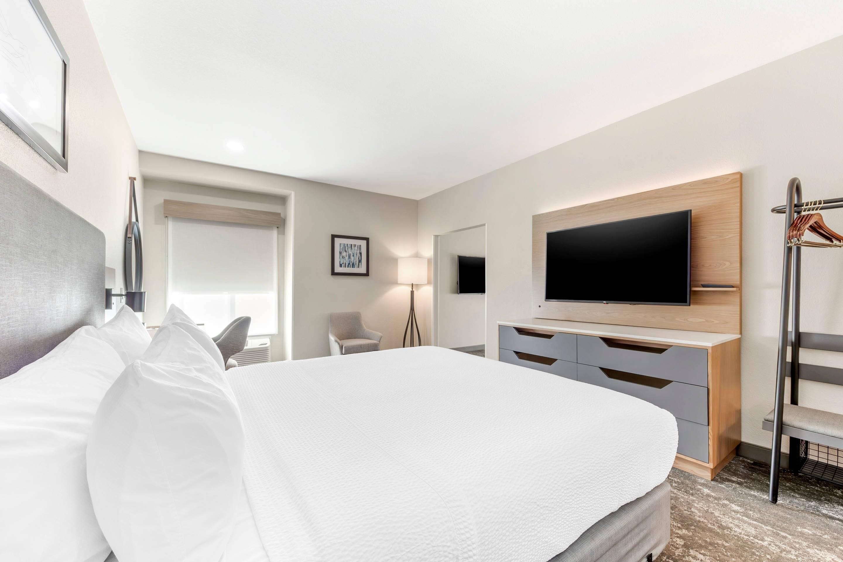 A modern hotel room with a large bed, a flat-screen TV mounted on a wooden panel, a desk with drawers, a chair, and a lamp.