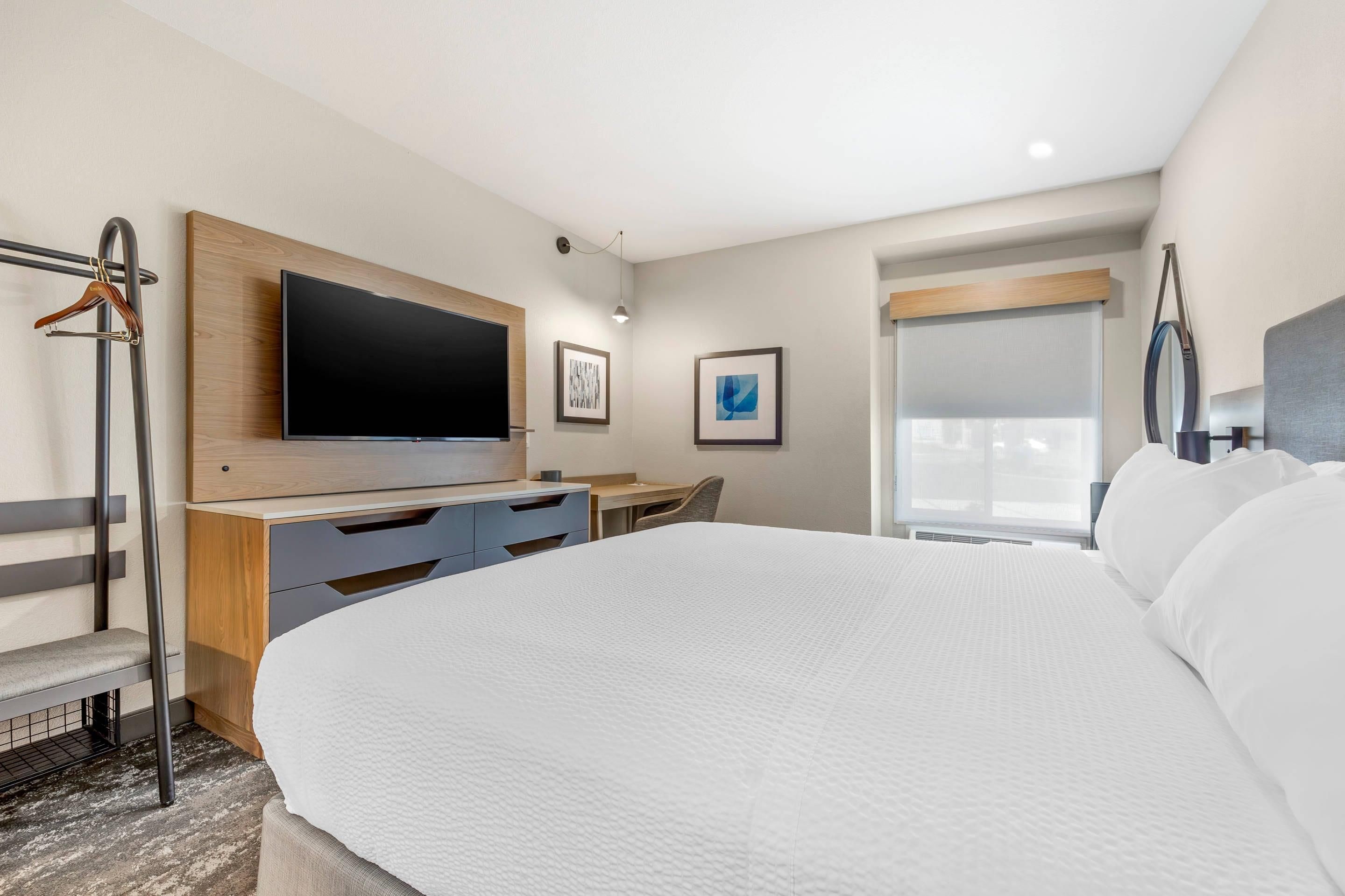 A modern hotel room with a large bed, a desk, a flat-screen TV, and a window with blinds.