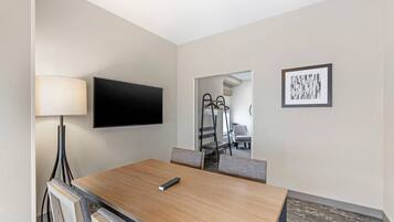 A hotel room with a wooden dining table, a flat-screen TV mounted on the wall, a floor lamp, a framed abstract artwork, and a view of the hallway and staircase.