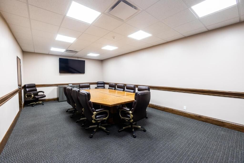 A conference room with a large rectangular table surrounded by 10 black office chairs, a carpeted floor, and a white wall with a mounted screen.