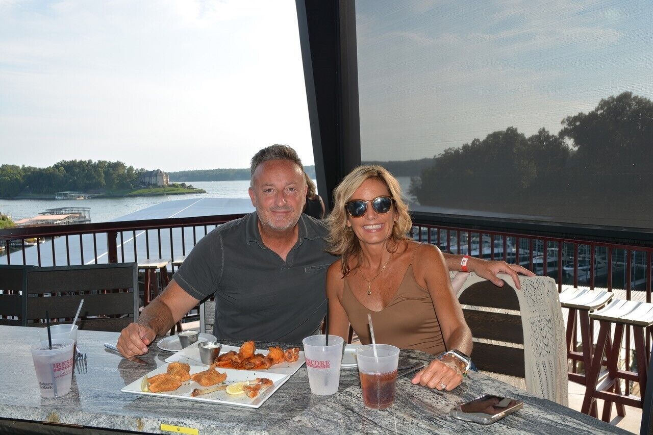 A couple dining at an outdoor table with a view of a lake and a boat.