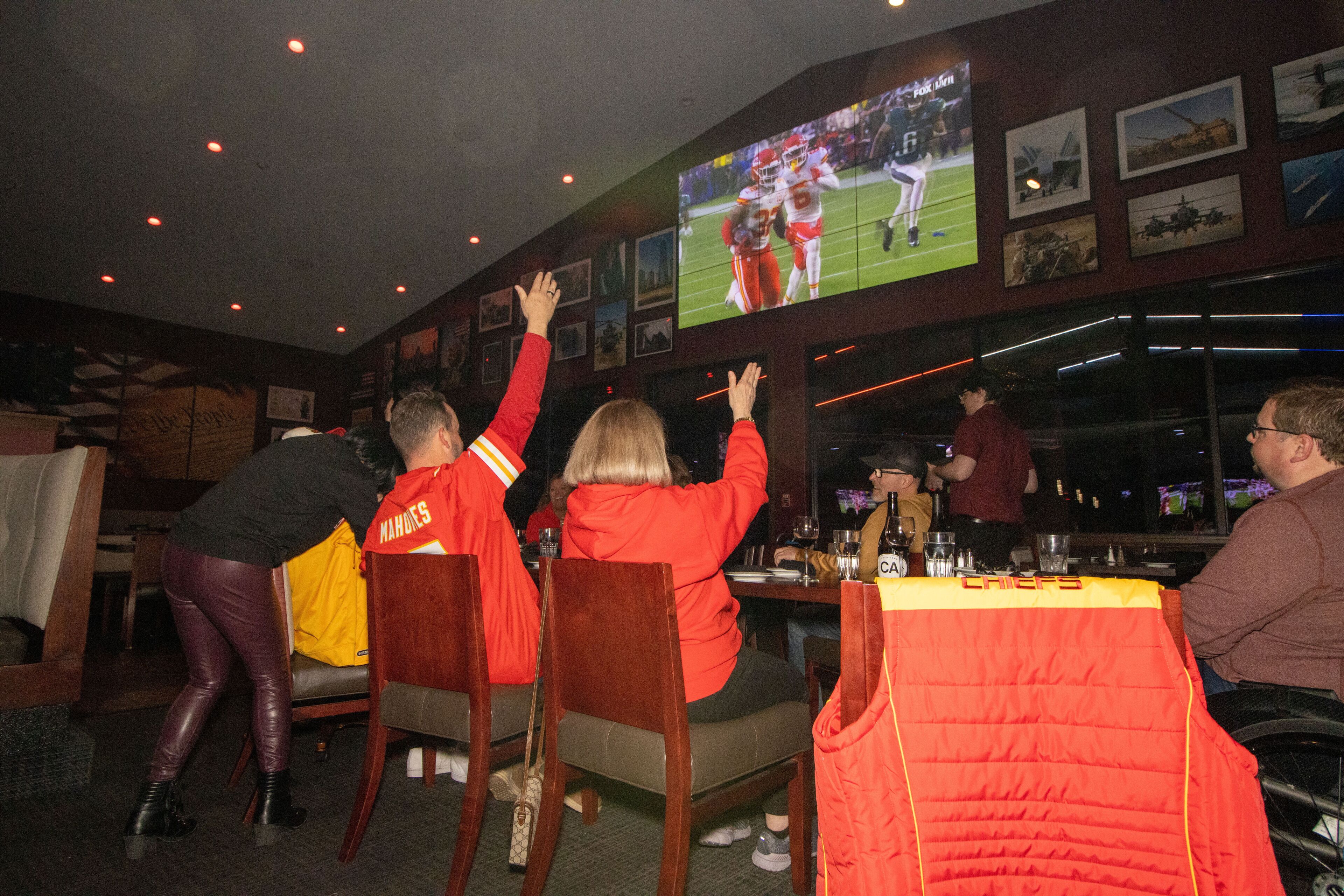 A group of people watching a football game on a large screen in a restaurant with wooden chairs and tables.