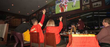 A group of people watching a football game on a large screen in a restaurant with wooden chairs and tables.