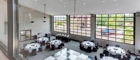 A spacious conference room with multiple tables and chairs, large windows with a view of the outside, and a ceiling with recessed lighting.
