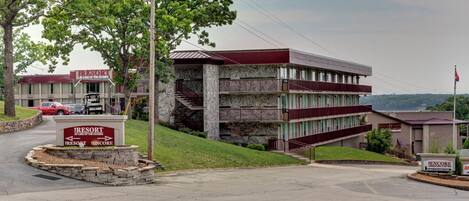 A multi-story building with a stone facade and a red roof, surrounded by greenery and a parking area.