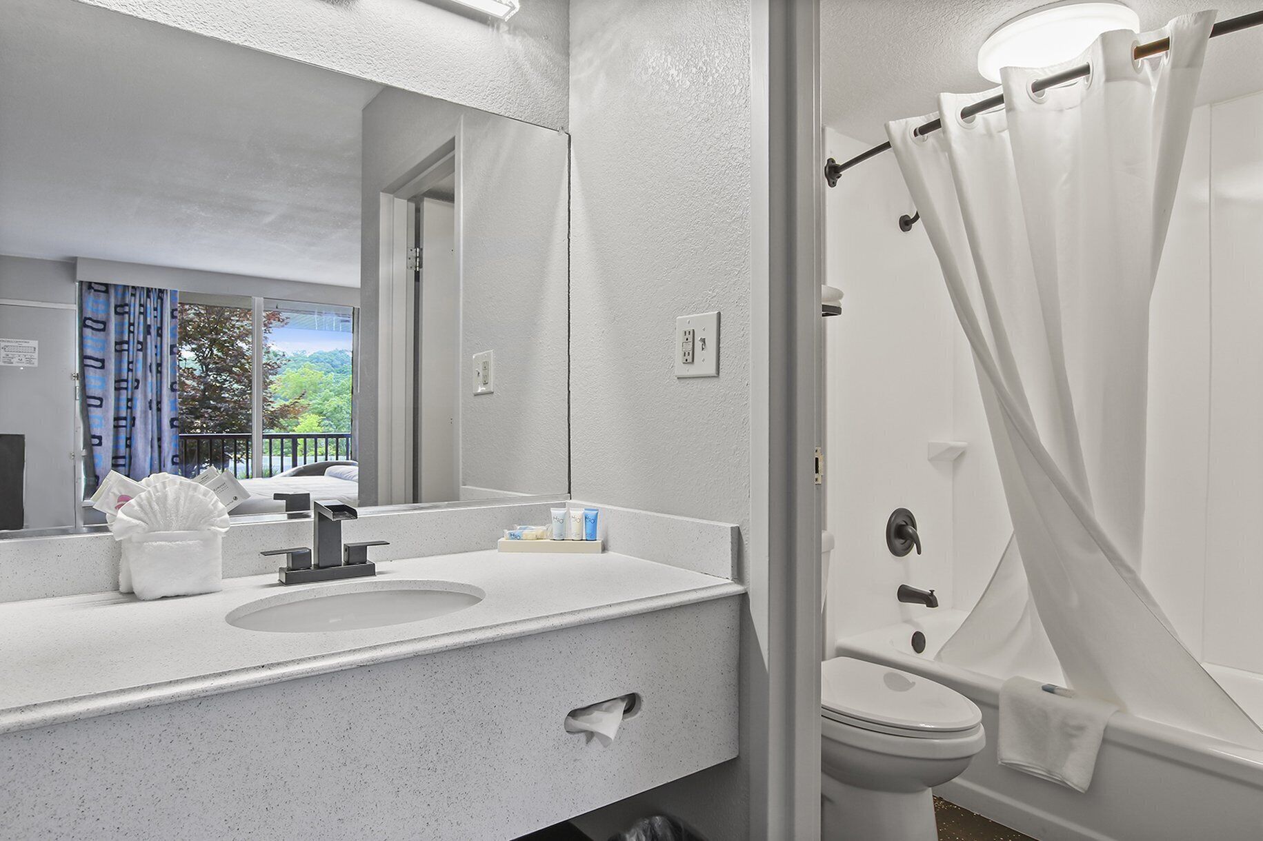 Une salle de bain avec un lavabo blanc, un miroir et des toilettes. Il y a un rideau de douche et une fenêtre donnant sur des arbres.