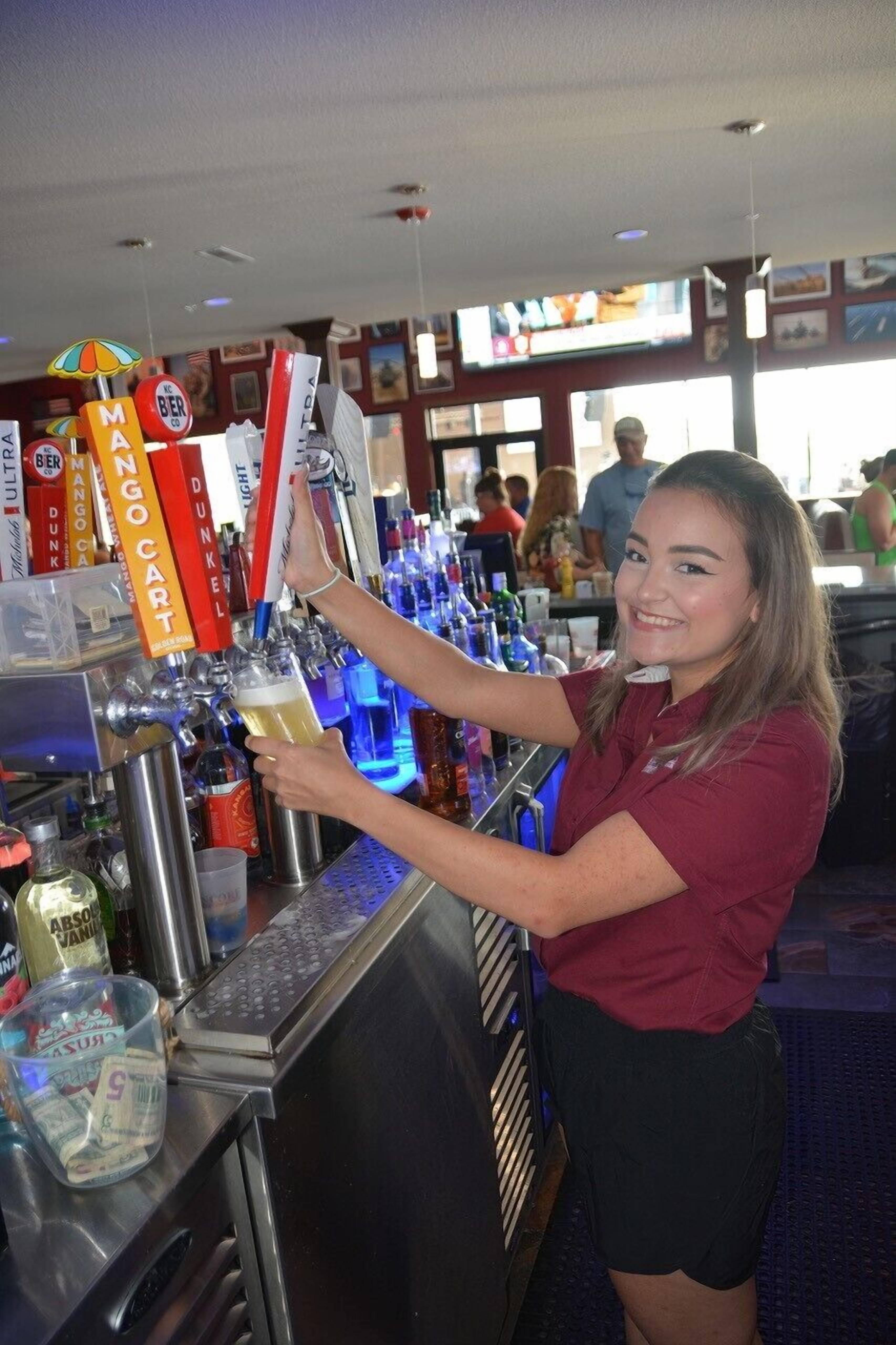 Un espace bar avec un barman préparant une boisson, une caisse enregistreuse, ainsi que diverses bouteilles et verres.