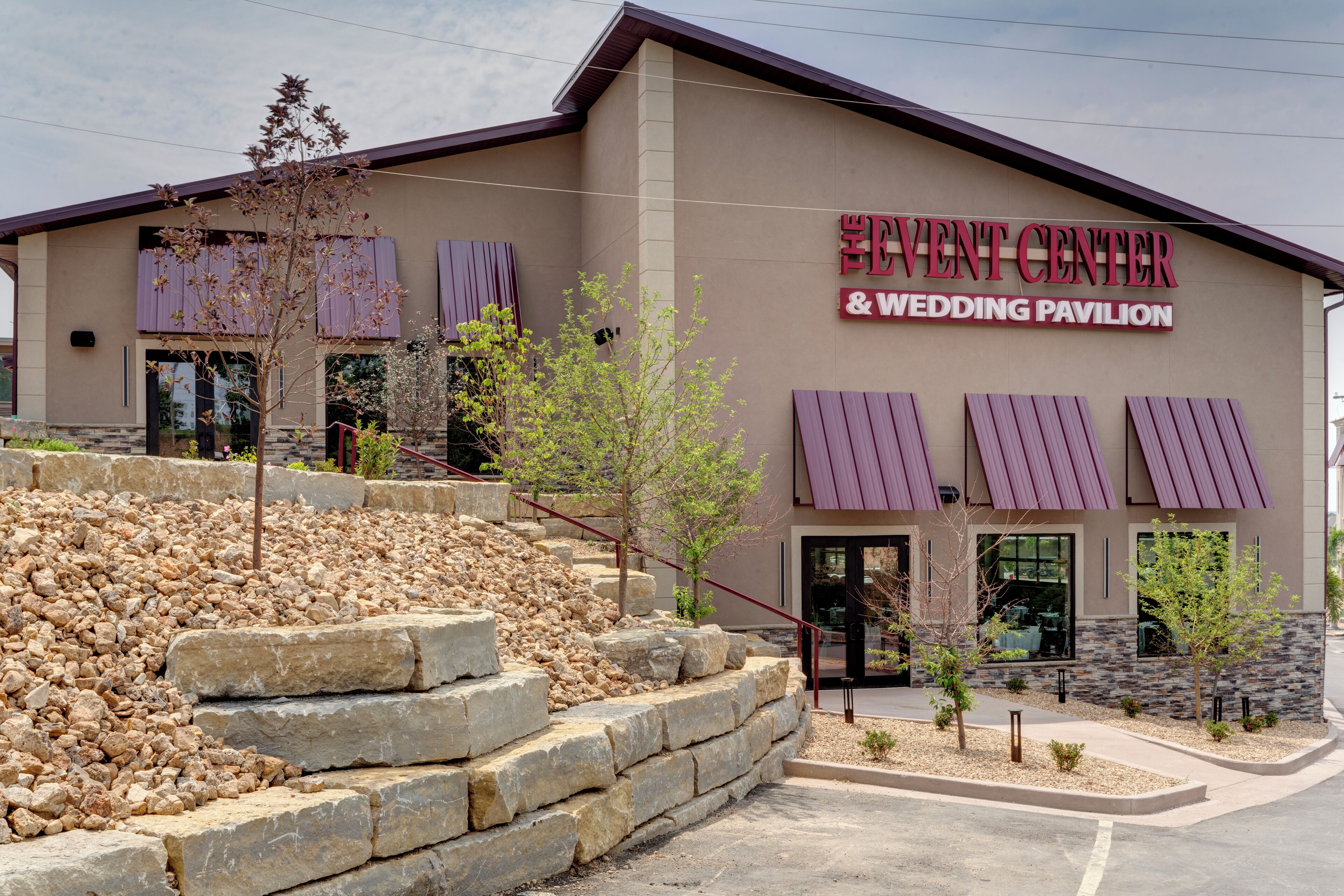 A building labeled "EVENT CENTER & WEDDING PAVILION" with a stone facade, purple awnings, and a landscaped entrance area.