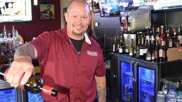 Un coin bar avec un barman qui verse une boisson dans des verres, un écran de télévision et un réfrigérateur rempli de bouteilles.