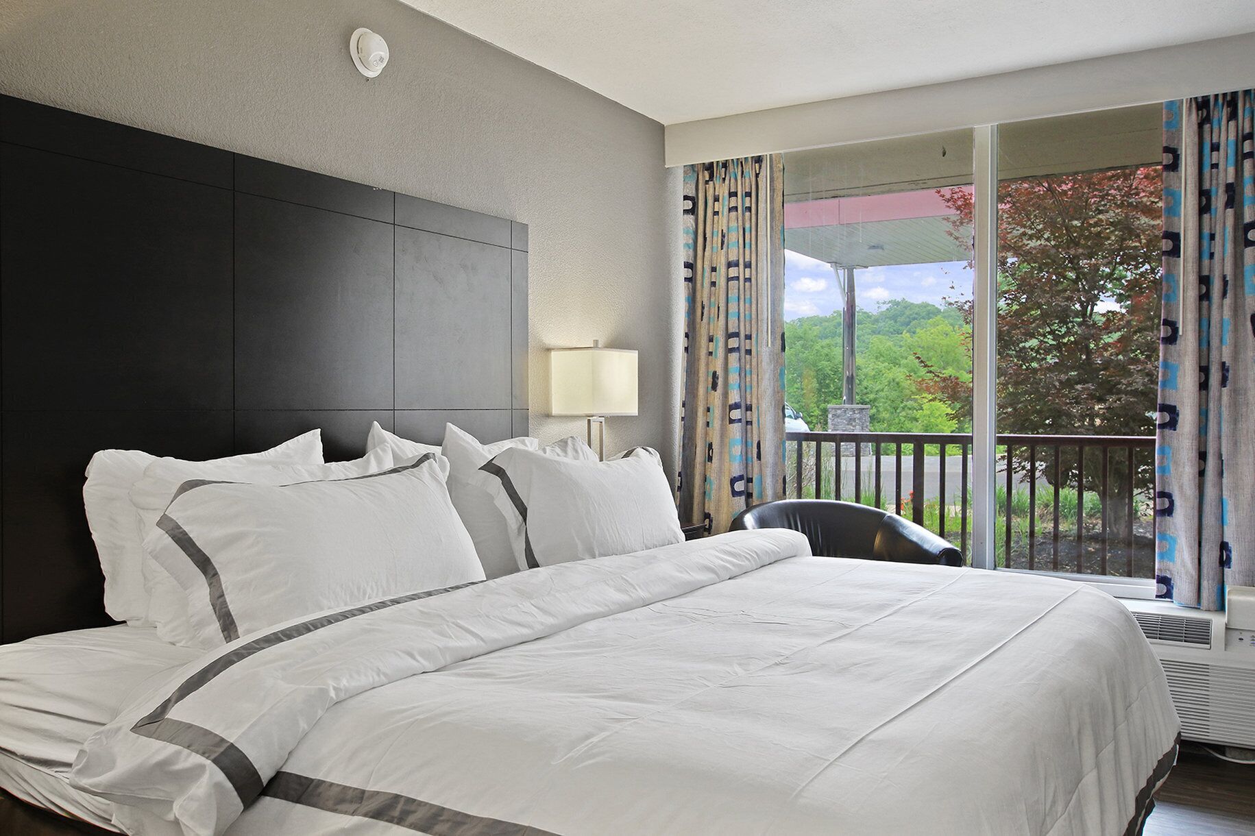 A hotel room with a large bed, a headboard, a nightstand, a lamp, and a view of the outdoors through a balcony with curtains.