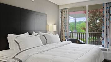 A hotel room with a large bed, a headboard, a nightstand, a lamp, and a view of the outdoors through a balcony with curtains.