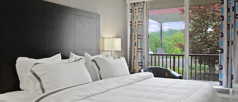 Une chambre d’hôtel avec un grand lit, une tête de lit, une table de chevet, une lampe et une vue sur l’extérieur grâce à un balcon agrémenté de rideaux.